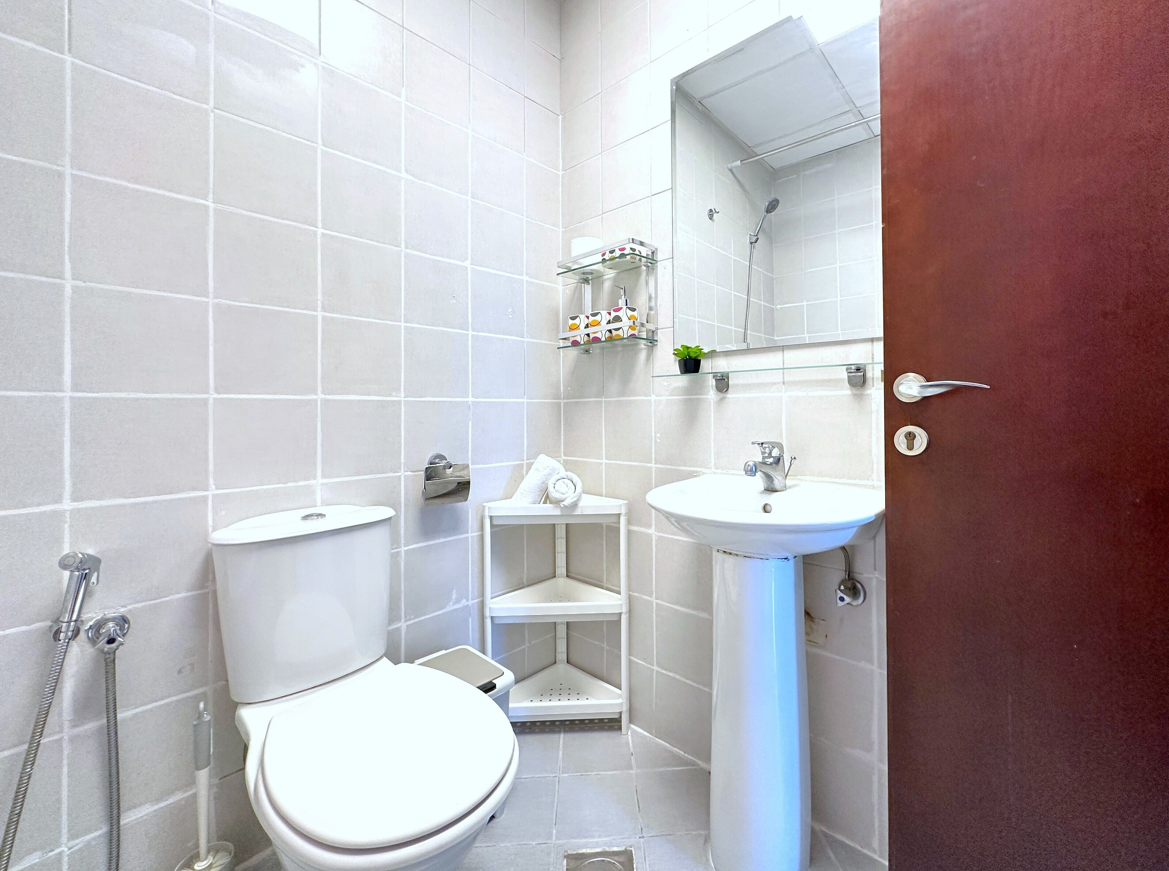 A bathroom with a toilet, sink, and shower. There is a mirror above the sink and a shelf with toiletries.