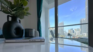 A modern room with a large window offering a city view, a black vase with green leaves, a candle, and a book on a glass table. - Luxury Brand New Studio  (Dubai)