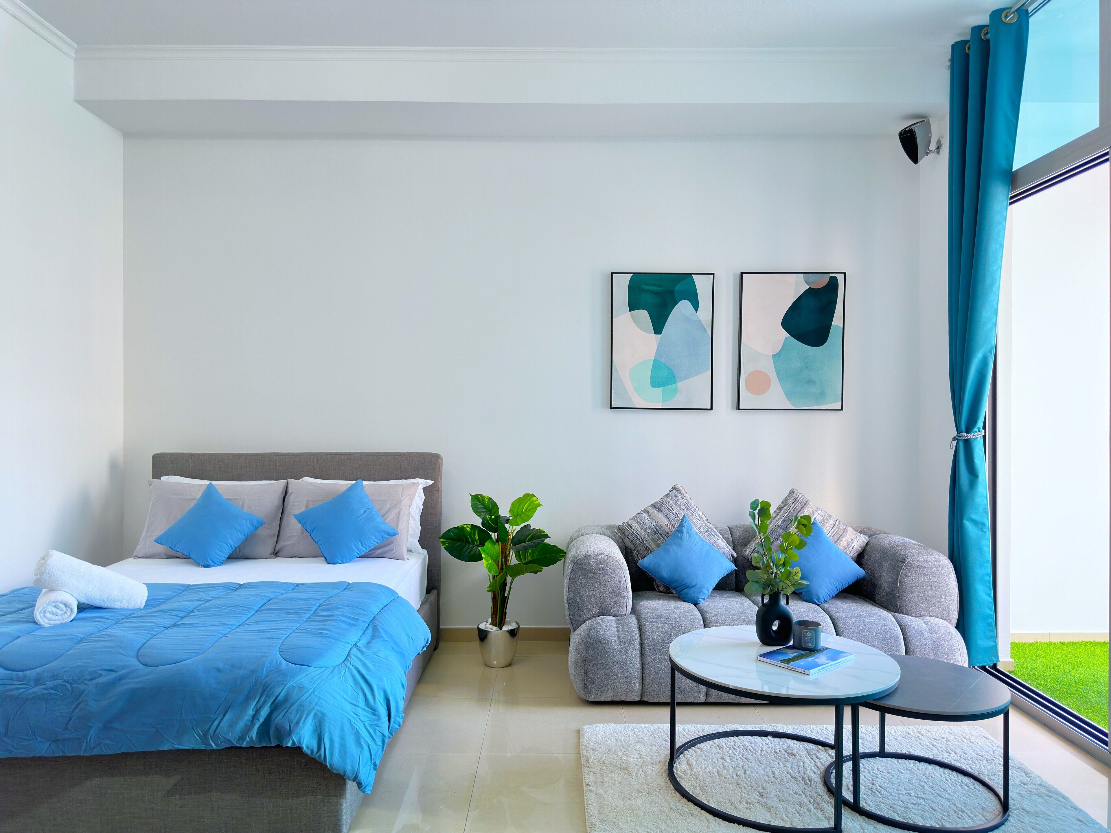Une chambre à coucher moderne avec un lit, un canapé gris, une petite table ronde et deux tableaux abstraits.