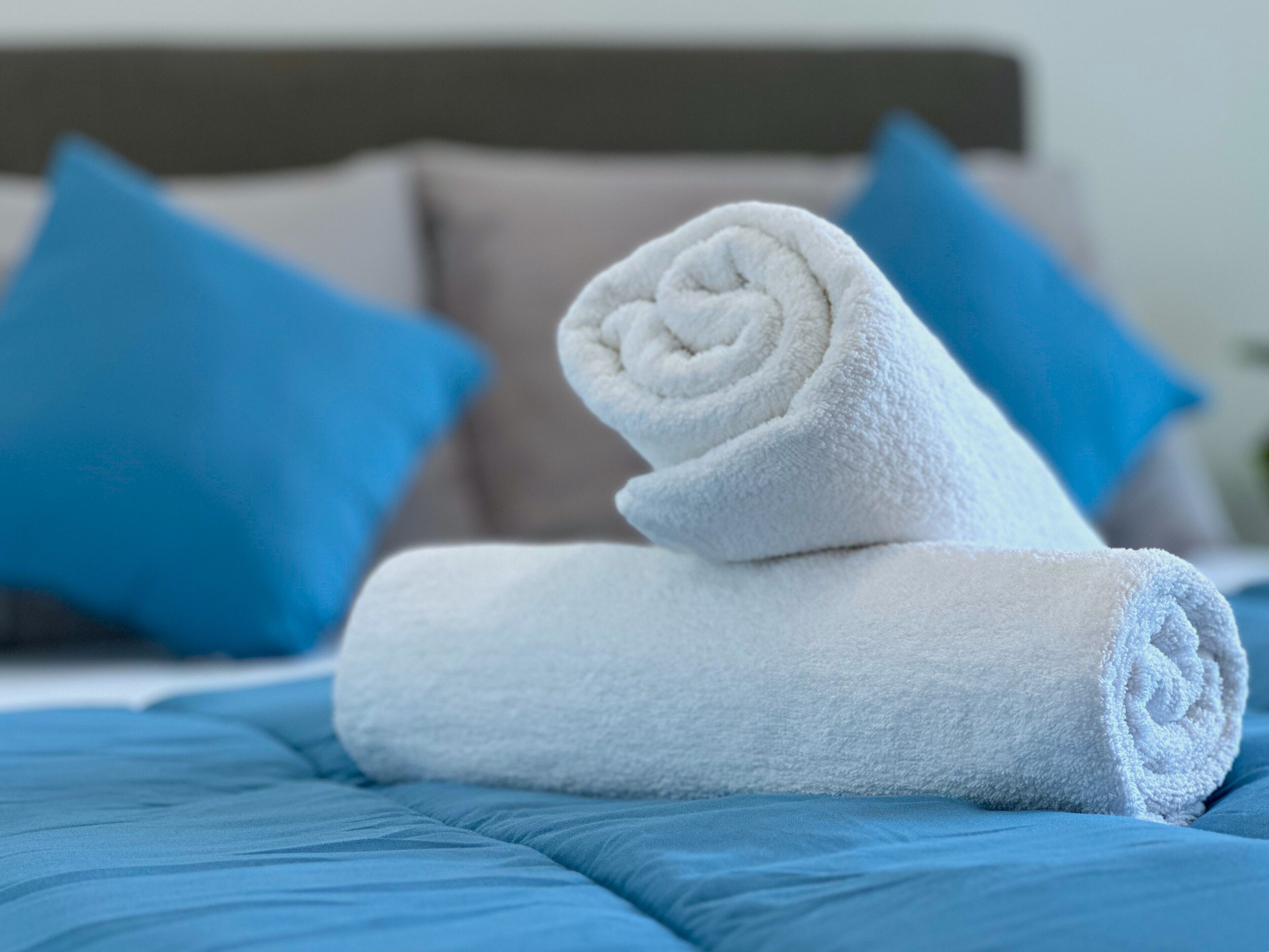 Ein ordentlich bezogenes Bett mit blauer Bettwäsche und am Fuß gerollten weißen Handtüchern.