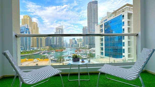 A balcony with two chairs, a small table, and a view of a cityscape with modern buildings and a marina. - Luxury Brand New Studio  (Dubai)