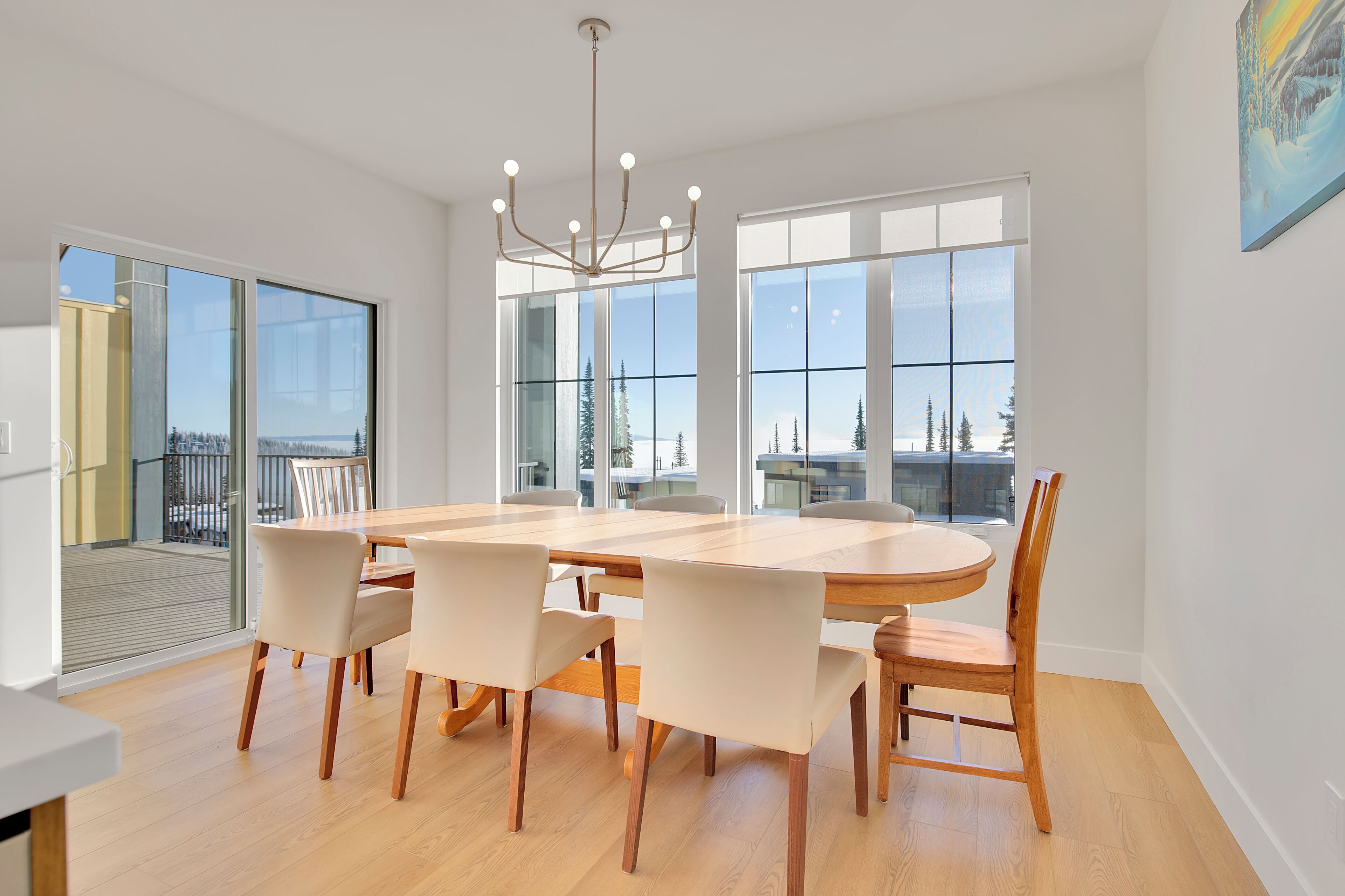 Un vaste espace dédié à la salle à manger, avec une grande table en bois et six chaises, de grandes fenêtres offrant une vue sur les arbres et les bâtiments, et un lustre moderne.