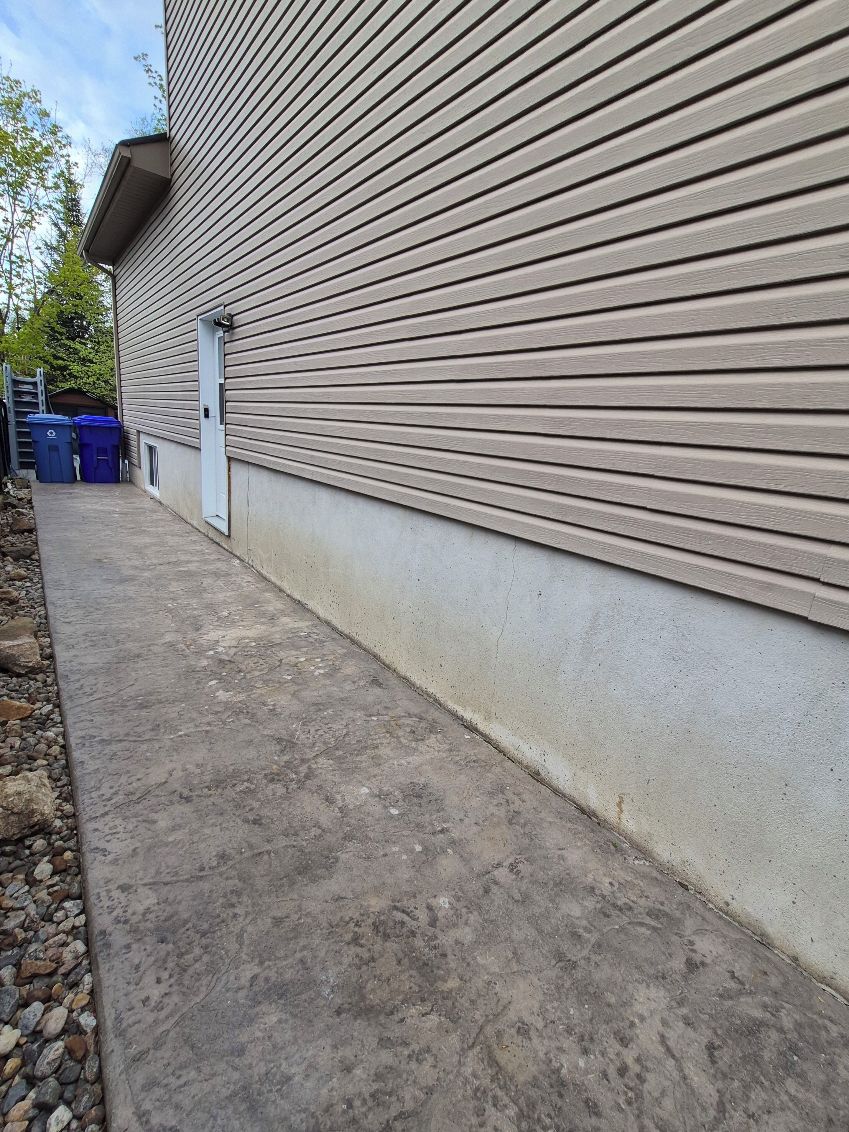 A building exterior with horizontal siding, a concrete pathway, and blue recycling bins.