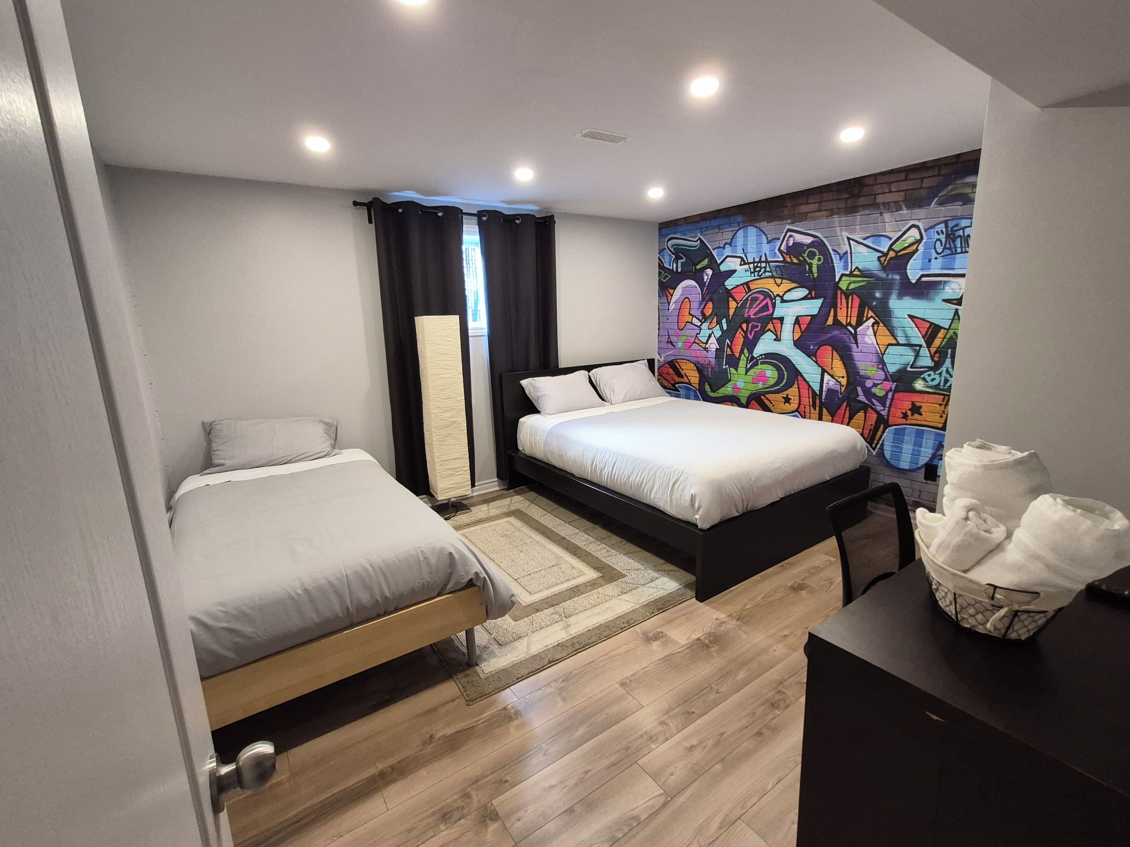 Un dormitorio con dos camas, un ventanal con cortinas, una pared con grafitis y un escritorio con una cesta.