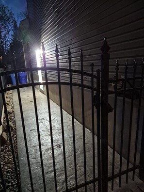A gated area with a concrete pathway, metal fence, and a building with horizontal siding. - Spacious 1-bedroom basement unit (full bathroom/shared laundry) (Gatineau)