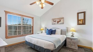 A bedroom with a large bed, a ceiling fan, a window with blinds, a nightstand with a lamp, and a wall-mounted mirror. - Blue Sky Lodge  For 4 (Branson West)