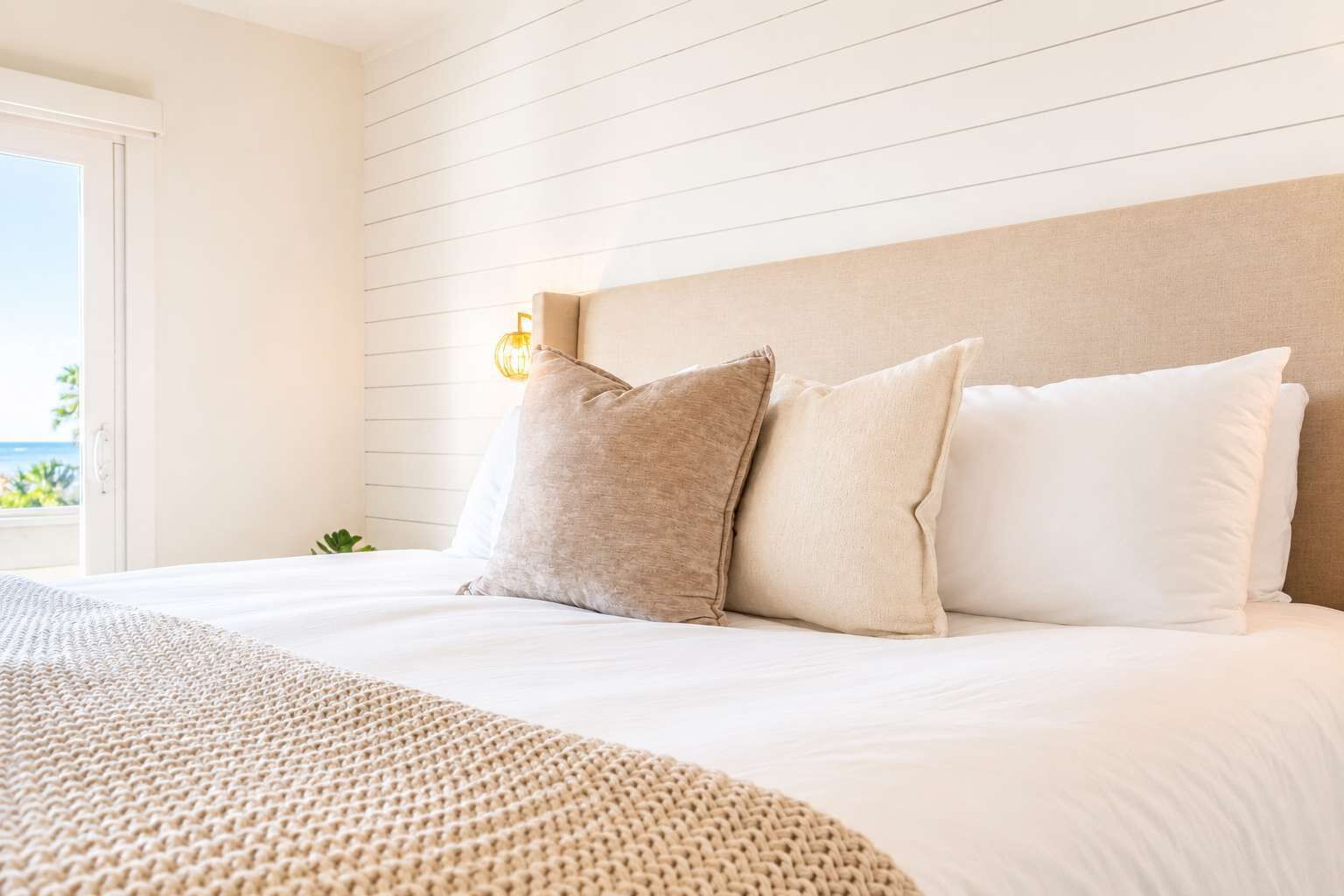 A bedroom with a bed, pillows, a bedside lamp, and a view of the outdoors.
