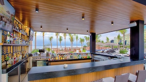 Eine Bar mit Blick auf Palmen und das Meer, ausgestattet mit einer Holzbalkendecke und einer Auswahl an Flaschen.