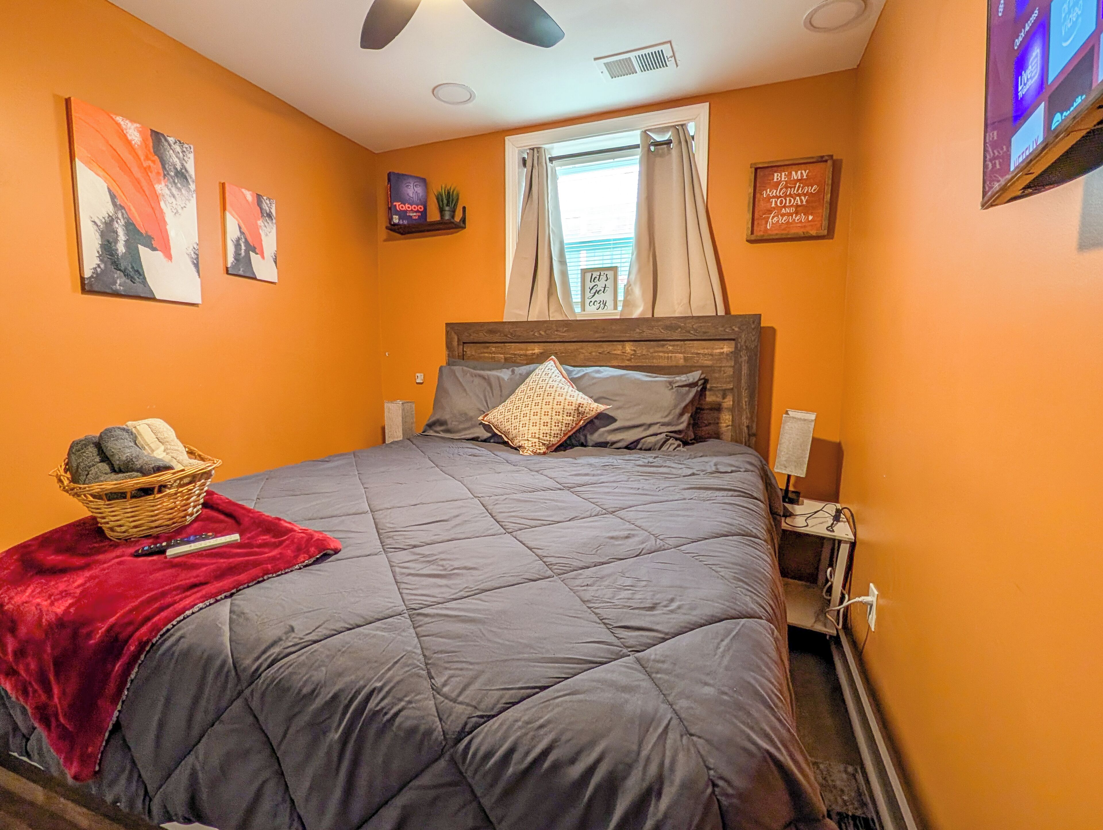 A bedroom with a bed, a red throw blanket, a wooden headboard, a nightstand, a wall-mounted TV, and a window with curtains.