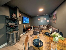 Una sala moderna con una pared gris oscuro, un televisor de pantalla plana, un sofá de cuero marrón, una mesa de comedor de madera con platos negros y una pequeña planta.
