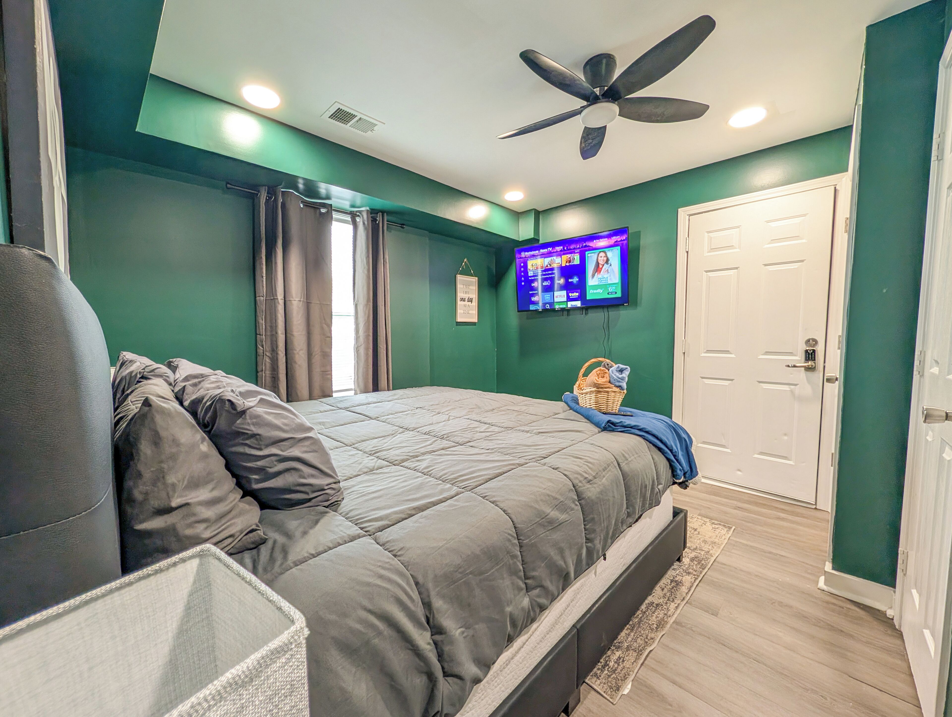 Une chambre à coucher avec un lit, un ventilateur de plafond, un téléviseur fixé au mur et une porte.