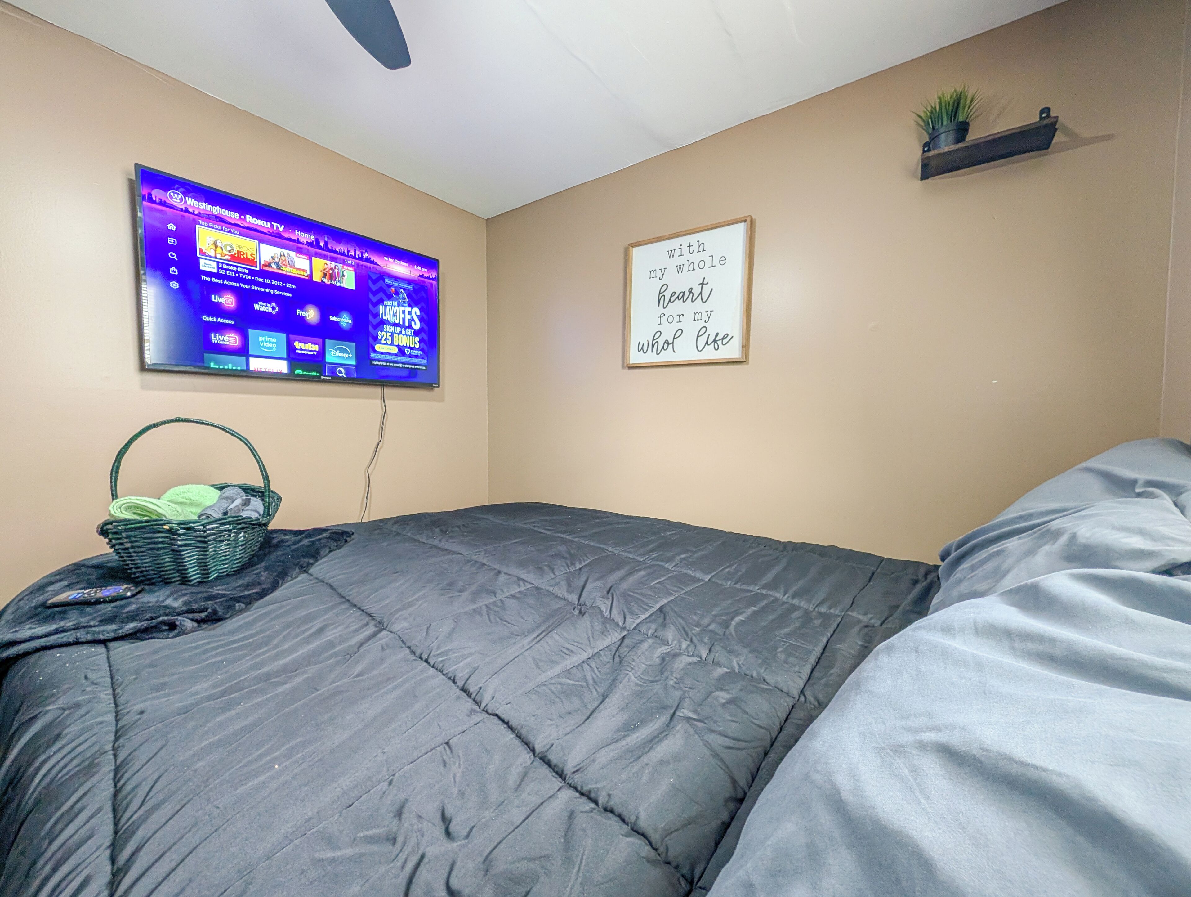 Un dormitorio con cama, televisor montado en la pared, estante con una planta y una frase motivacional.