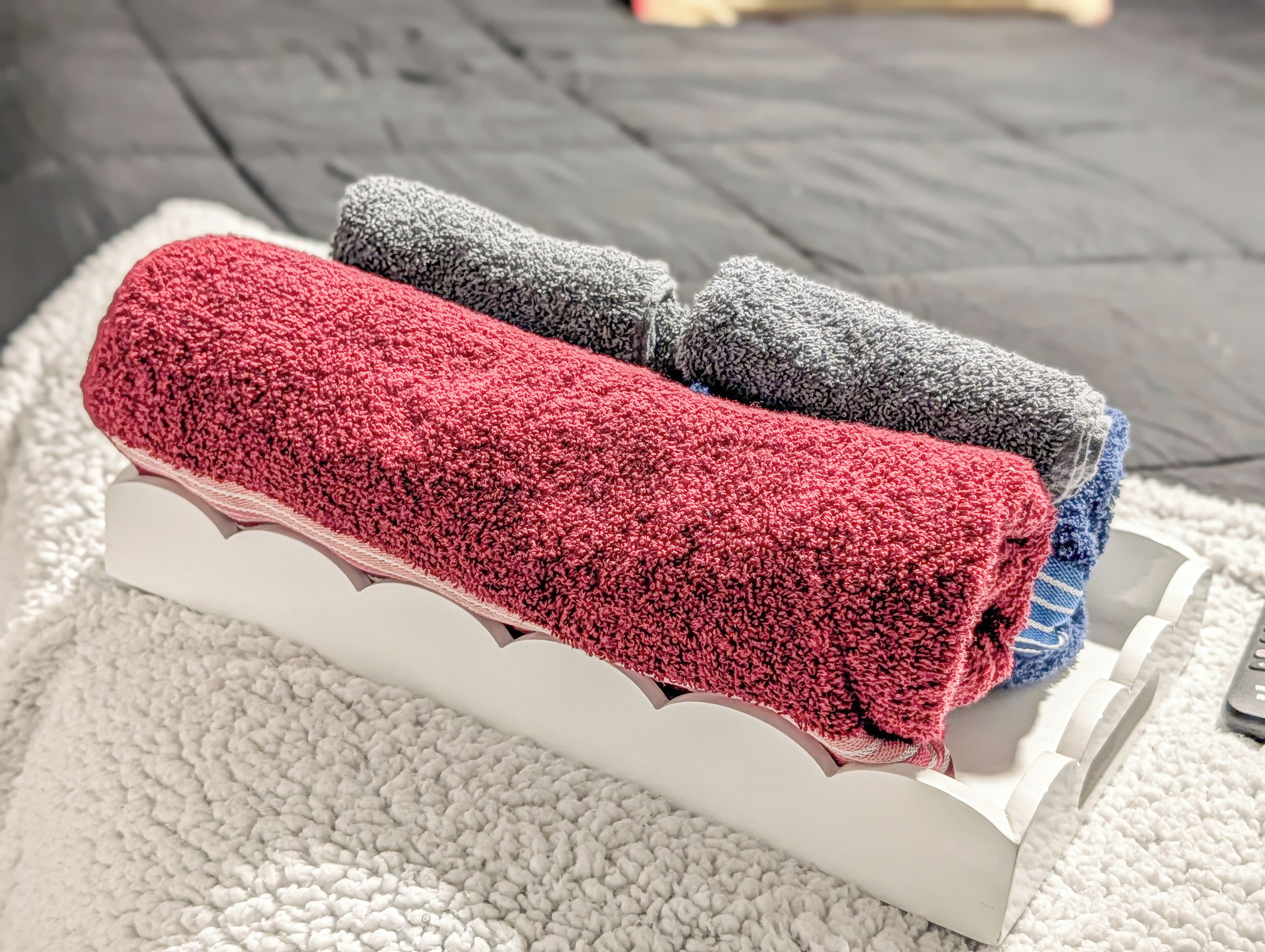 A white, scalloped-edge tray holding rolled towels in red, gray, and blue colors.