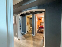 Un pasillo con piso de madera, un baño con ducha y un dormitorio con cama y una colcha roja.