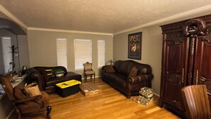 A living room with a dark brown leather sofa, a wooden coffee table, and a wooden cabinet. There are three windows with blinds, a painting on the wall, and a wooden floor. - Spacious 4-bedroom house with AC in fantastic Houston neighborhood (Houston)
