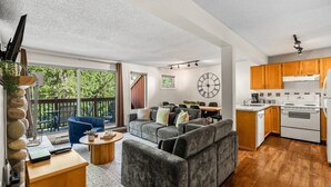 Een moderne woonkamer met een grijze bank, een eethoek en een keuken met houten kastjes en witte apparaten.