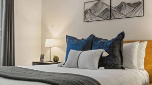 Een opgemaakt bed met een grijze deken, een houten hoofdbord en meerdere kussens in blauw- en wit tinten.