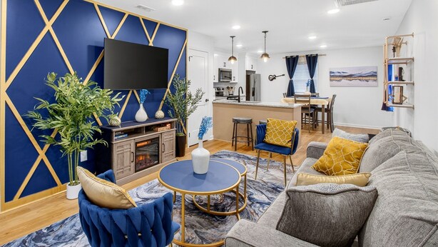 A modern living room with a blue and gold accent wall, a flat-screen TV, a fireplace, and a dining area with a kitchen in the background. - MusicCityRetreat|3BR2BA|Arcade|Sleep8|EasyDTCommute (Madison)