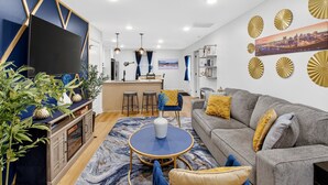 A modern living room with a grey sofa, a round coffee table, and a TV mounted on a blue wall. There is a kitchen area with stools and a bar counter. - MusicCityRetreat|3BR2BA|Arcade|Sleep8|EasyDTCommute (Madison)