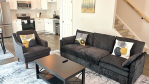 Ein modernes Wohnzimmer mit grauem Sofa, einem passenden Sessel und einem Couchtisch. Im Hintergrund ist eine Küche mit weißen Schränken und Edelstahlgeräten zu sehen.