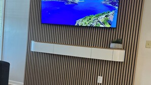 Een moderne kamer met een grote televisie bevestigd aan een houten paneelwand, een glazen tafel met een decoratieve witte schaal, en een klein plantje op een plank onder de televisie.