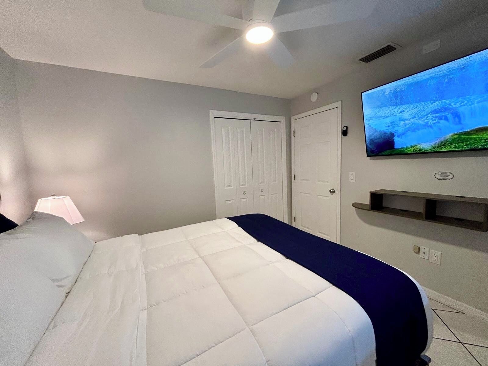 A bedroom with a bed, a ceiling fan, a TV on the wall, and a door.