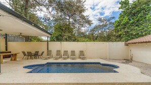 Ein Garten mit Schwimmbad, Liegestühlen, einem Tisch und einer überdachten Sitzgruppe.