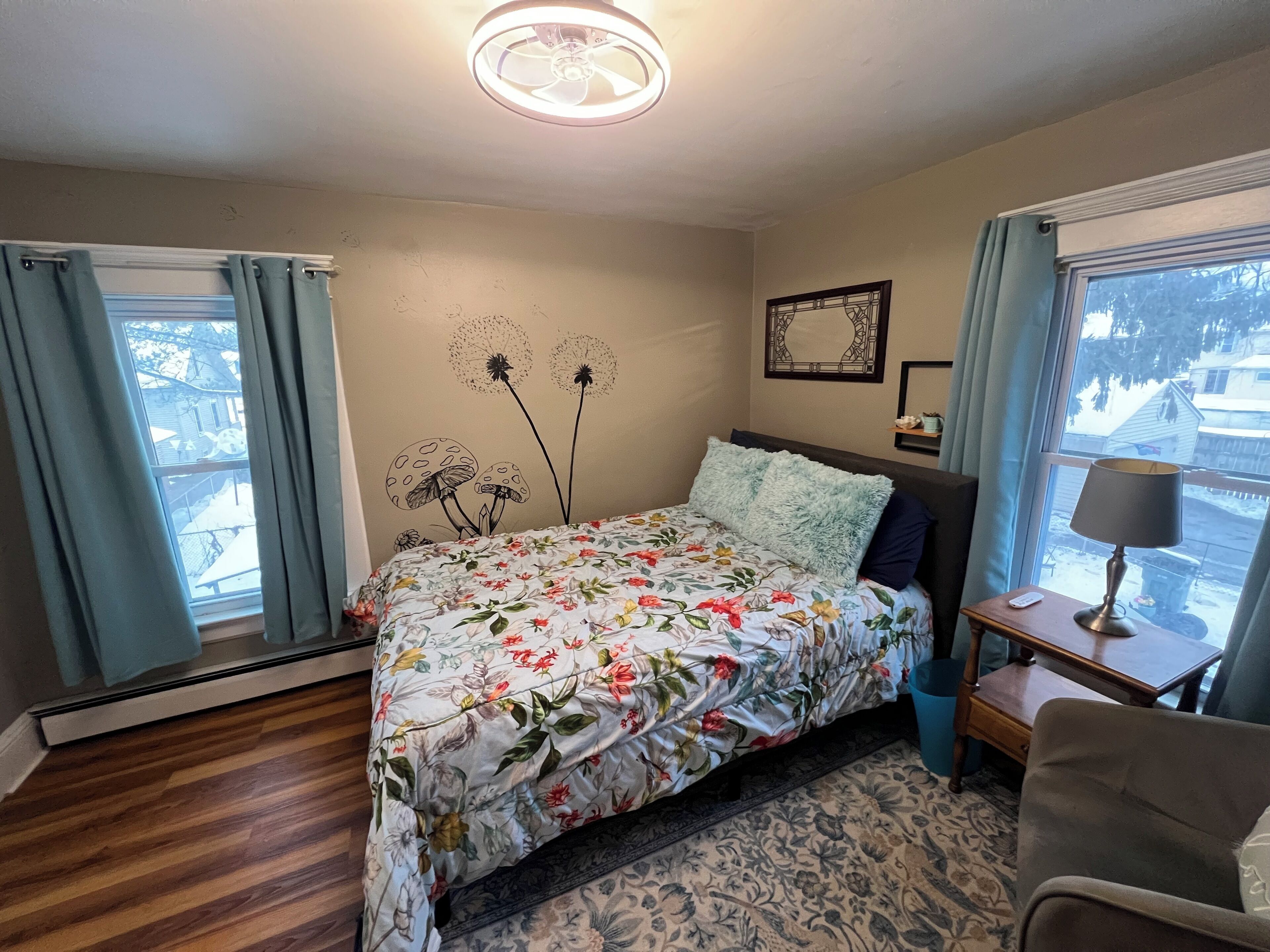 A bedroom with a floral bedspread, a ceiling fan, a bedside table with a lamp, and a view of the outside through a window.