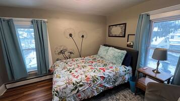 A bedroom with a floral bedspread, a ceiling fan, a bedside table with a lamp, and a view of the outside through a window.