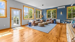Un salon spacieux doté de grandes fenêtres, d’un plancher en bois et d’un coin salon confortable avec un canapé et des poufs.