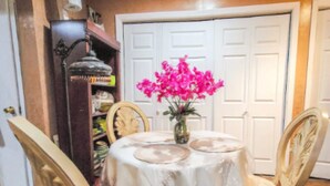 A dining area with a round table covered by a white tablecloth, surrounded by four chairs with cushioned seats. A vase with pink flowers is placed on the table.