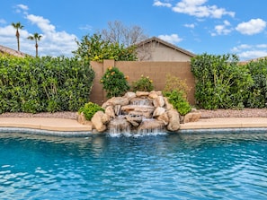 A swimming pool with a rock waterfall feature, surrounded by well-maintained hedges and palm trees. - Private Pool Oasis Near Spring Training & Luke AFB (Surprise)
