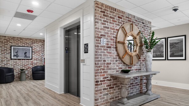 A reception area with a brick wall, a round wooden clock, a white reception desk, and a framed picture on the wall. - Cozy Condo with Rooftop Views (Bay St. Louis)