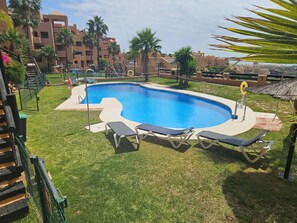 Una piscina amb gandules, una zona enjardinada i un edifici al fons.