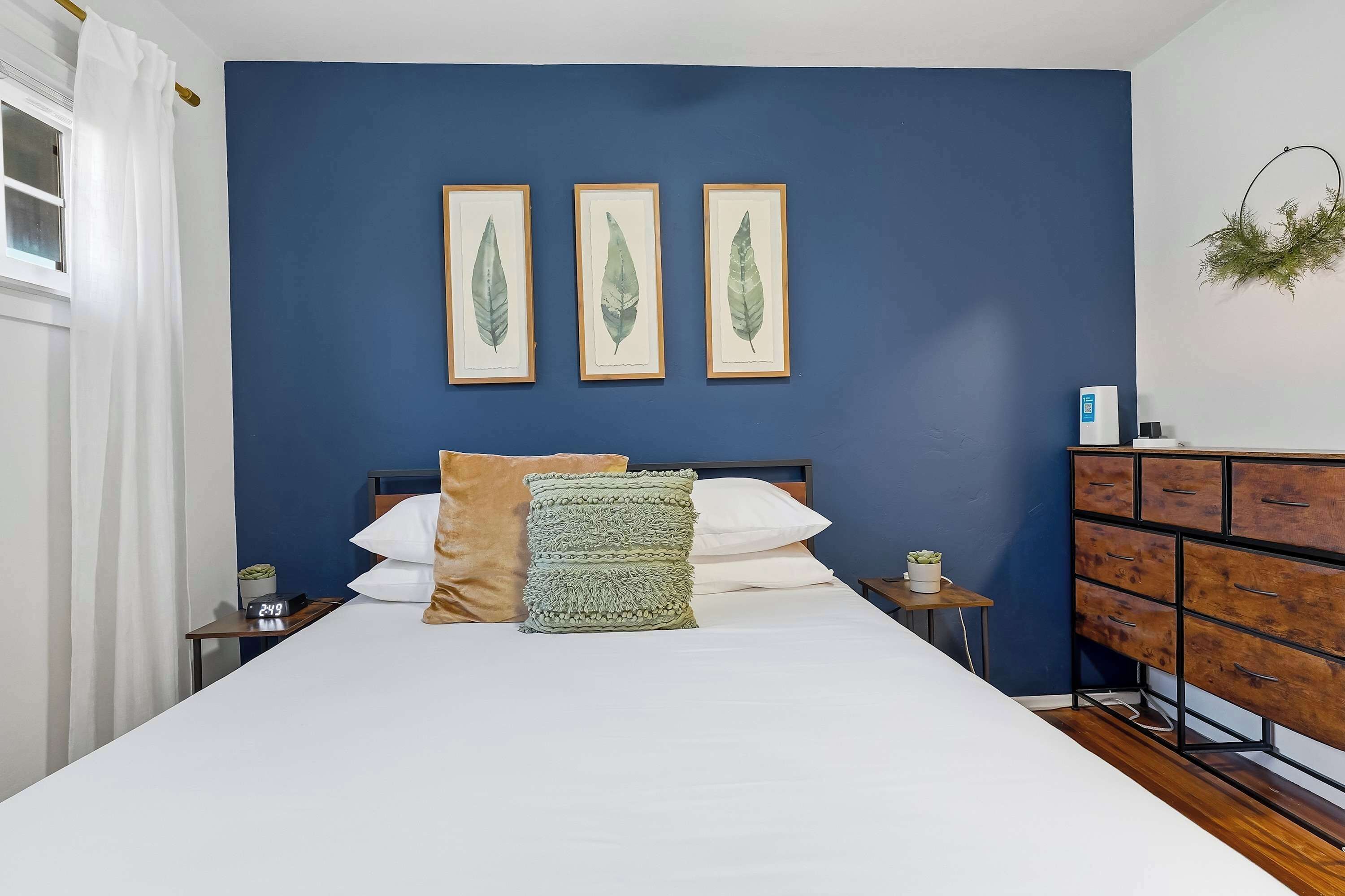 A bedroom with a large bed, a nightstand with a clock, a dresser, and three framed leaf artworks on the wall.