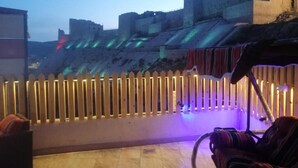 Une terrasse offrant une vue sur un fort historique, une clôture en bois et un espace détente agrémenté d’un coussin à rayures.