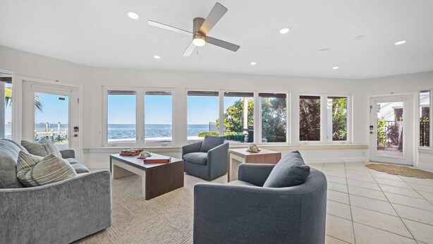 A spacious living room with a ceiling fan, large windows with ocean views, and a tiled floor. - Infinite Views! Heated Pool! Directly on Tampa Bay (Holmes Beach)