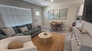 Uma sala de estar moderna com um sofá cinzento, uma poltrona branca, uma mesa de centro redonda e uma área de jantar com mesa e cadeiras.