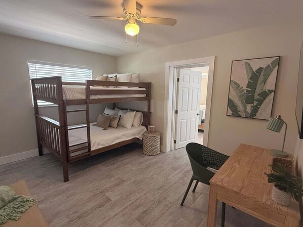 Une chambre avec un lit superposé, un bureau avec une chaise, un ventilateur de plafond et une porte donnant sur une autre pièce.