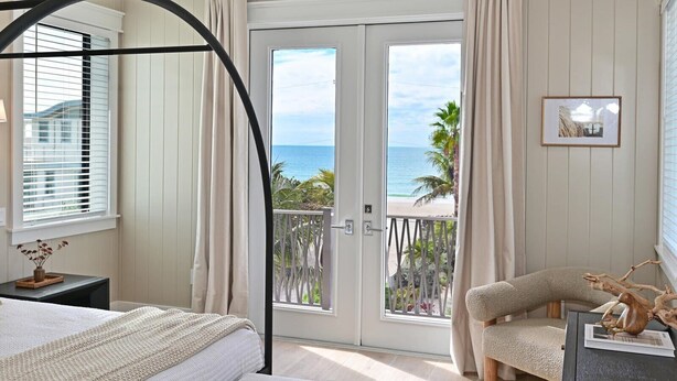 Una camera da letto con un letto, una sedia, un tavolino, una finestra con tende, una porta che dà su un balcone e un quadro appeso al muro.