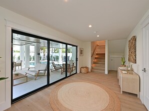 A spacious living area with a large sliding glass door leading to an outdoor patio with a pool, a staircase, and a small side table with decorative items. - Nirvana Sunset~Across from Beach, Heated Pool/Spa! (Holmes Beach)