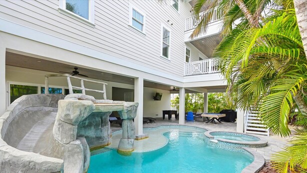 A pool with a waterfall feature, surrounded by a covered patio area with seating and a view of a white building. - Atlantis Beach House~Water Slide, Heated Pool/Spa! (Bradenton Beach)