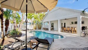A swimming pool with lounge chairs and umbrellas, surrounded by palm trees and a white building. - Birdsong-Steps to Bean Point Beach Heated Pool! (Anna Maria)