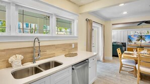 A modern kitchen with a double sink, wooden backsplash, and a dining area with a table and chairs. - Birdsong-Steps to Bean Point Beach Heated Pool! (Anna Maria)