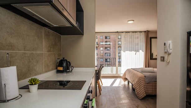 Een kleine hotelkamer met kitchenette, een bed met gestreepte beddengoed, uitzicht op een gebouw door het raam, en een telefoon aan de muur.
