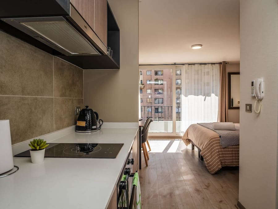 Quarto de hotel compacto com cozinha compacta, uma cama com roupa de cama listrada, vista de um edifício pela janela e um telefone na parede.