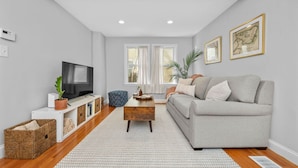 A modern living room with a grey sofa, a wooden coffee table, a TV on a white console, and a large window with white curtains. - Boston Condo with Free Parking (Boston)