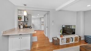 A modern kitchen with a marble countertop, white cabinetry, and a built-in oven. There is a dining area with a wooden table and chairs, and a living room with a flat-screen TV and a sofa. - Boston Condo with Free Parking (Boston)