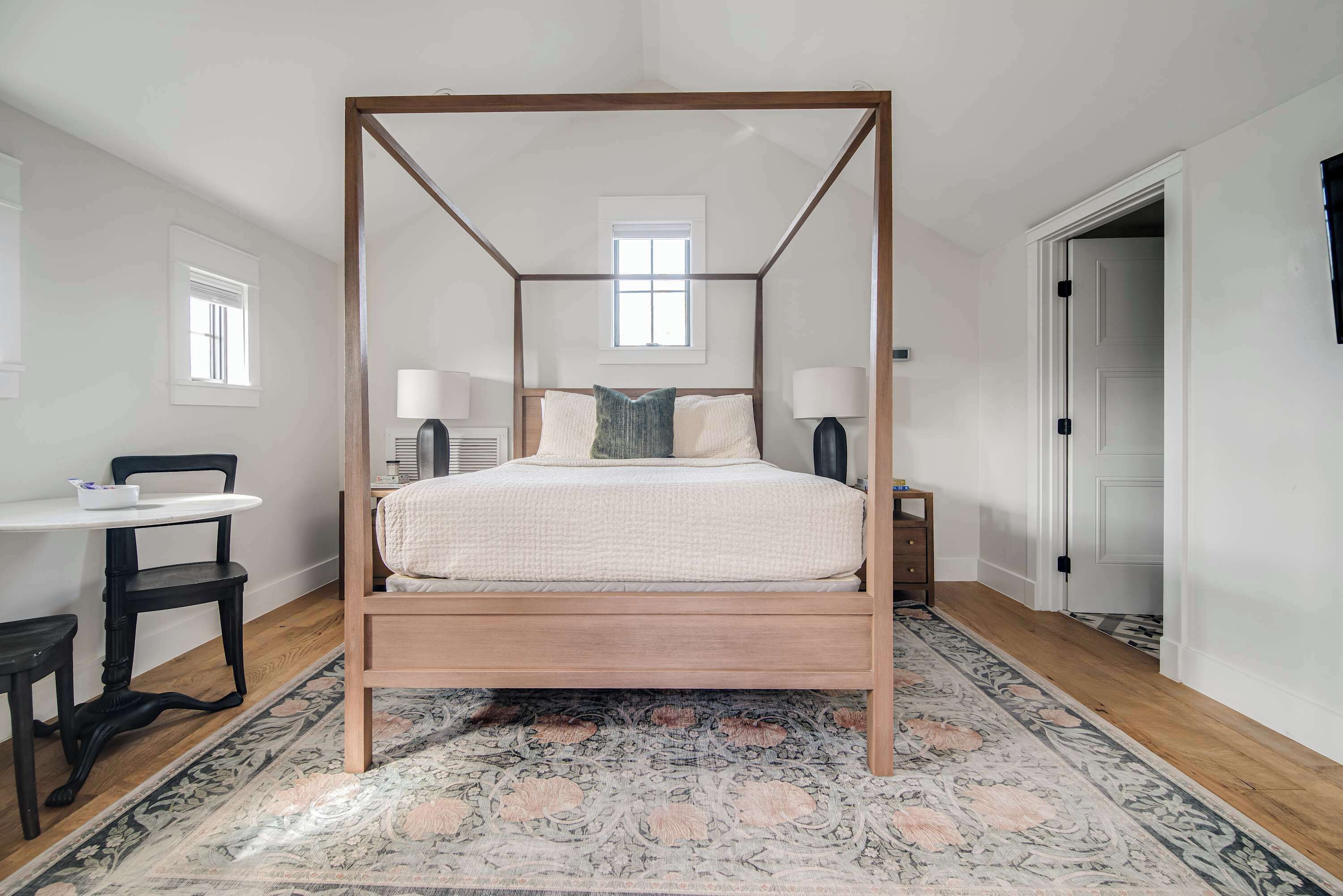 Ein Bett mit Baldachin, dessen Gestell aus Holz gefertigt ist, mit weißer Bettdecke und zwei Kissen. Zur Linken des Bettes steht ein kleiner Tisch mit einem Stuhl, zur Rechten ein Nachttisch mit einer Lampe. Ein gemustertes Teppichabdeckung bedeckt den Boden.