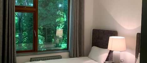 A hotel room with two beds, a window with curtains, a nightstand with a lamp, and a ceiling light.